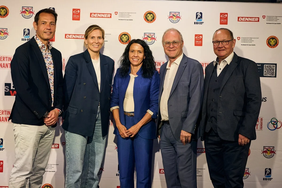 Five individuals stand side by side in front of a backdrop with various logos, including Red Bull, Swiss, Dennner, and others. They are dressed in formal and business casual attire, smiling at the camera.