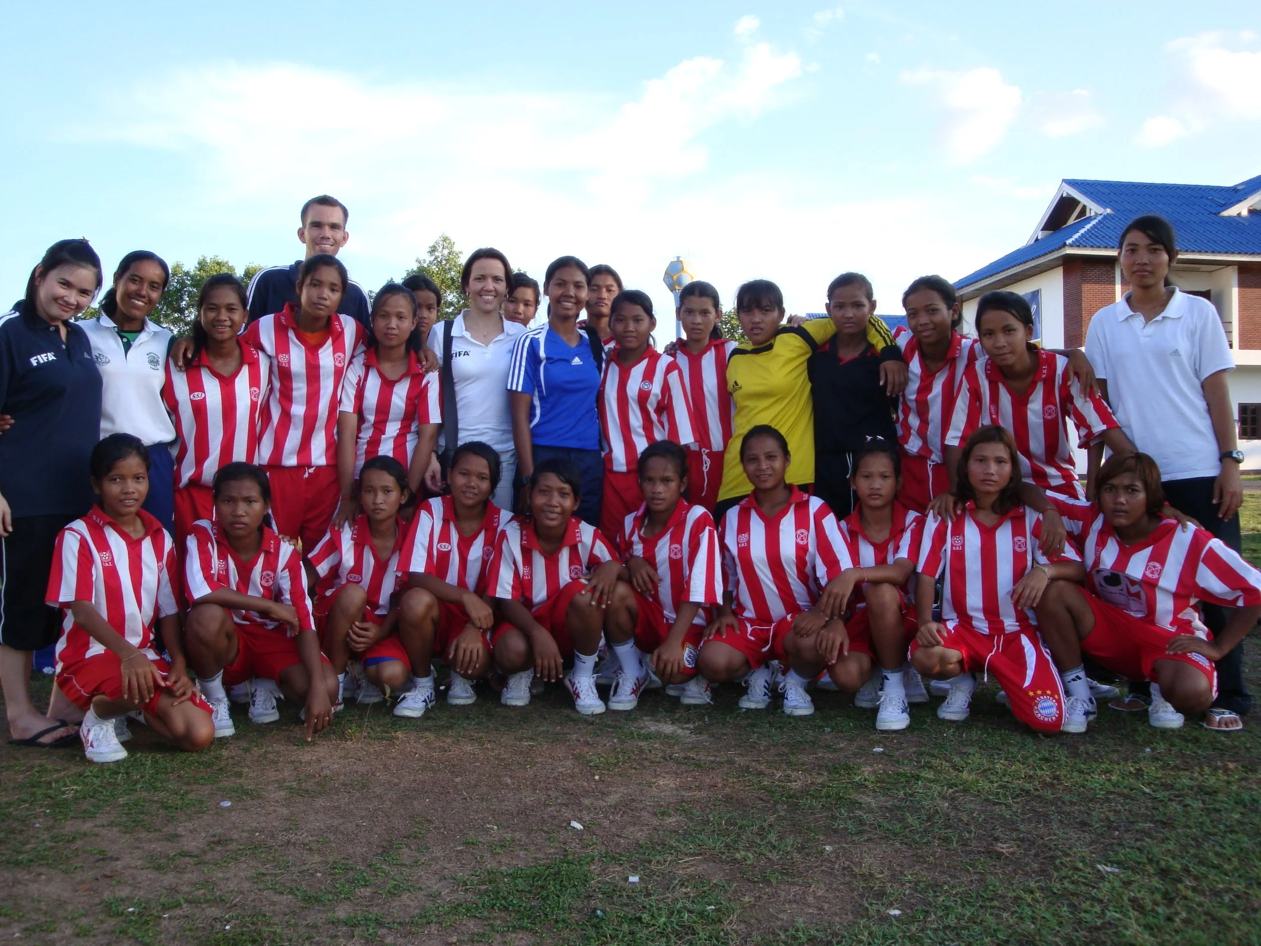 Group of young women and girls, some wearing sporting uniforms, standing outdoors in front of a building with a blue roof. They are smiling and posing for the photo.