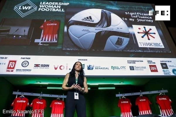 Woman speaking on stage at a sports leadership event, with red sports jerseys hanging behind her and a large screen displaying FIFA and Adidas logos, a football helmet, and various sponsor logos.