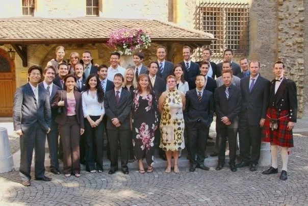 Group of 20 people in business attire standing outdoors in front of a rustic building with cobblestone pavement and a flower basket, for a formal or professional event.