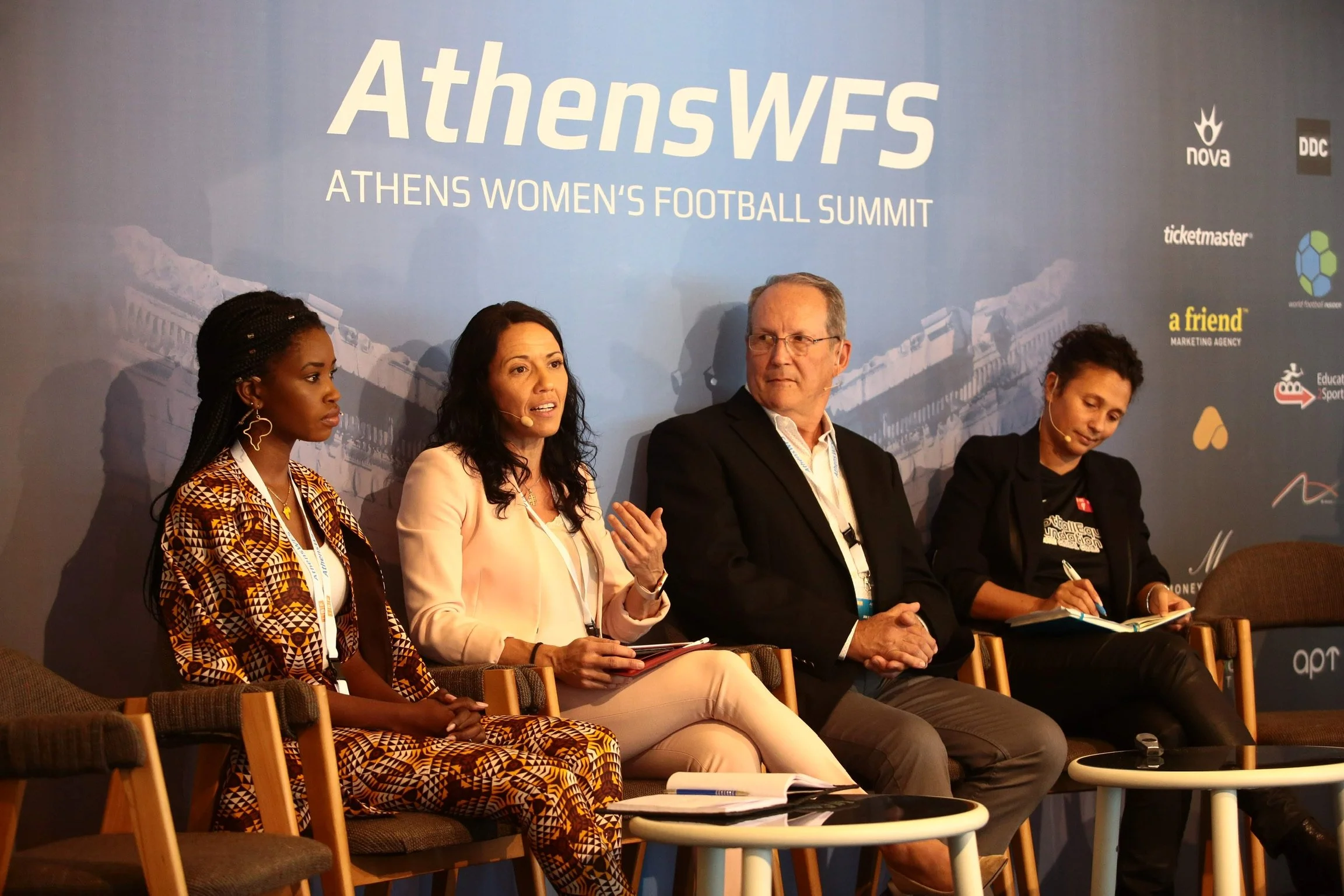 Four people sitting on chairs at the Athens Women's Football Summit, with a blue backdrop behind them displaying the event's title and logos of sponsors.