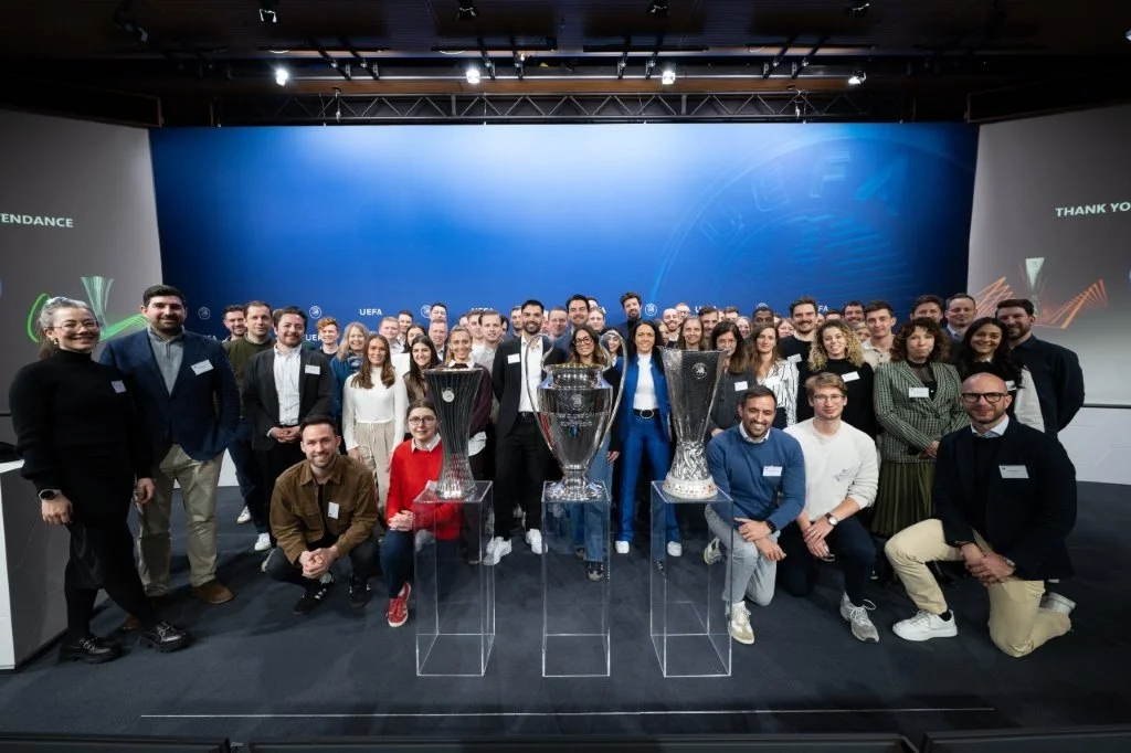 Group of people at a UEFA awards event with three large trophy sculptures on pedestals in front, smiling and posing for a photo in an indoor venue with blue and white background and presentation screens.