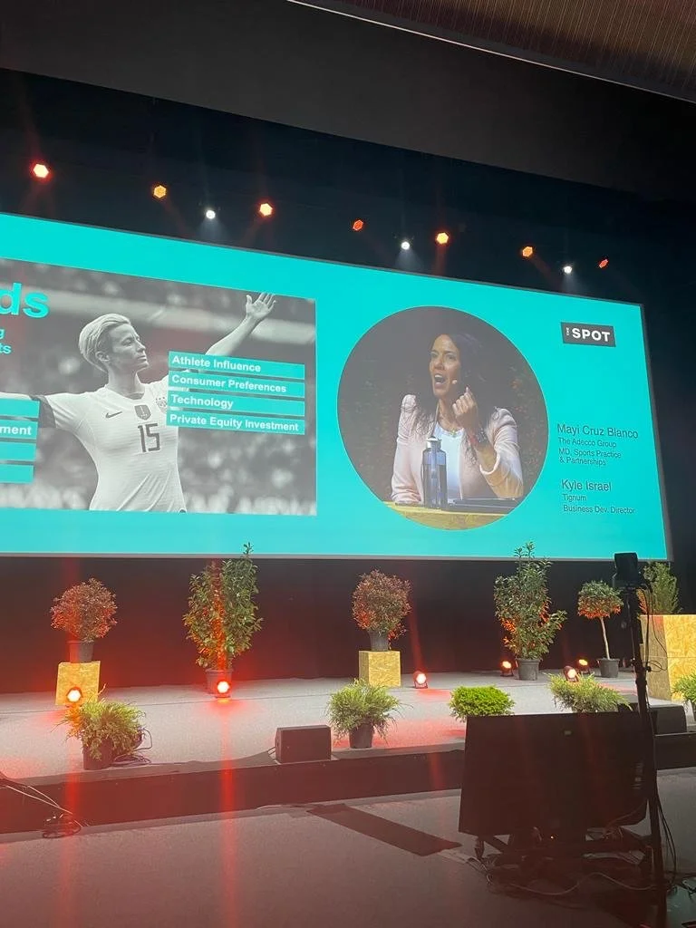 Large presentation screen showing a woman speaking at a conference, with a black and white photo of a female soccer player on the left and a circular photo of the speaker on the right. The stage has potted plants and lighting.