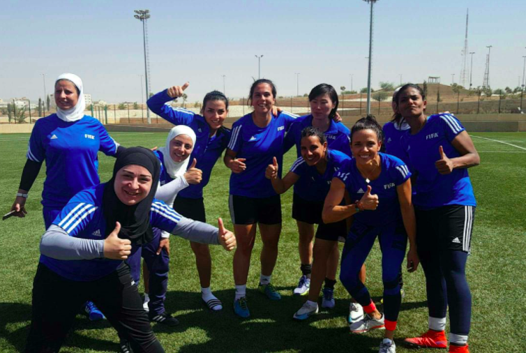 Group of female soccer players in blue jerseys posing on a soccer field.
