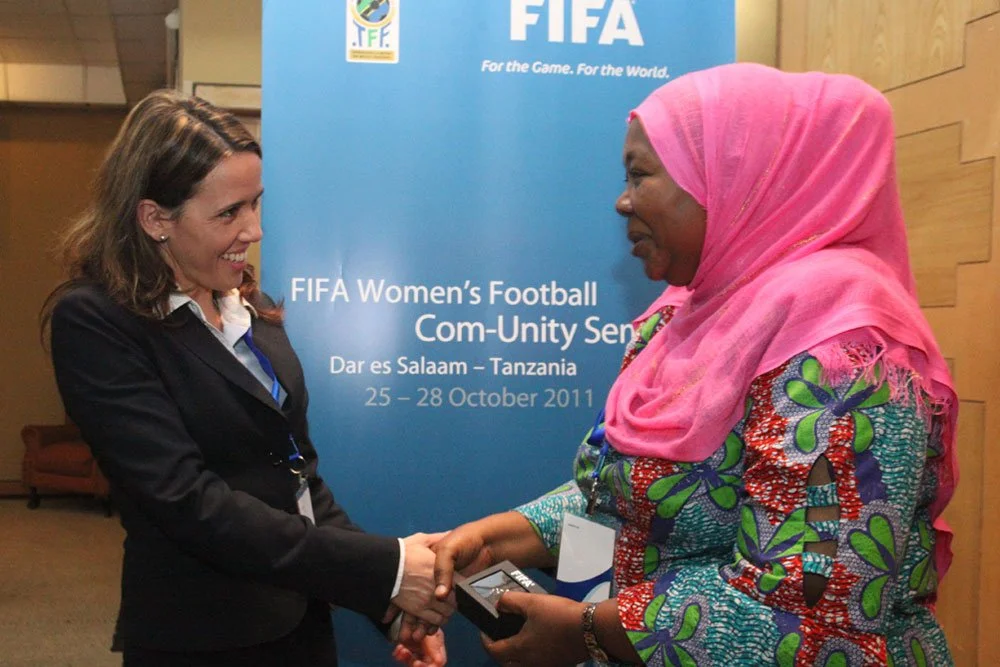 Four women standing together in front of FIFA banners, smiling, indoors at a professional event.