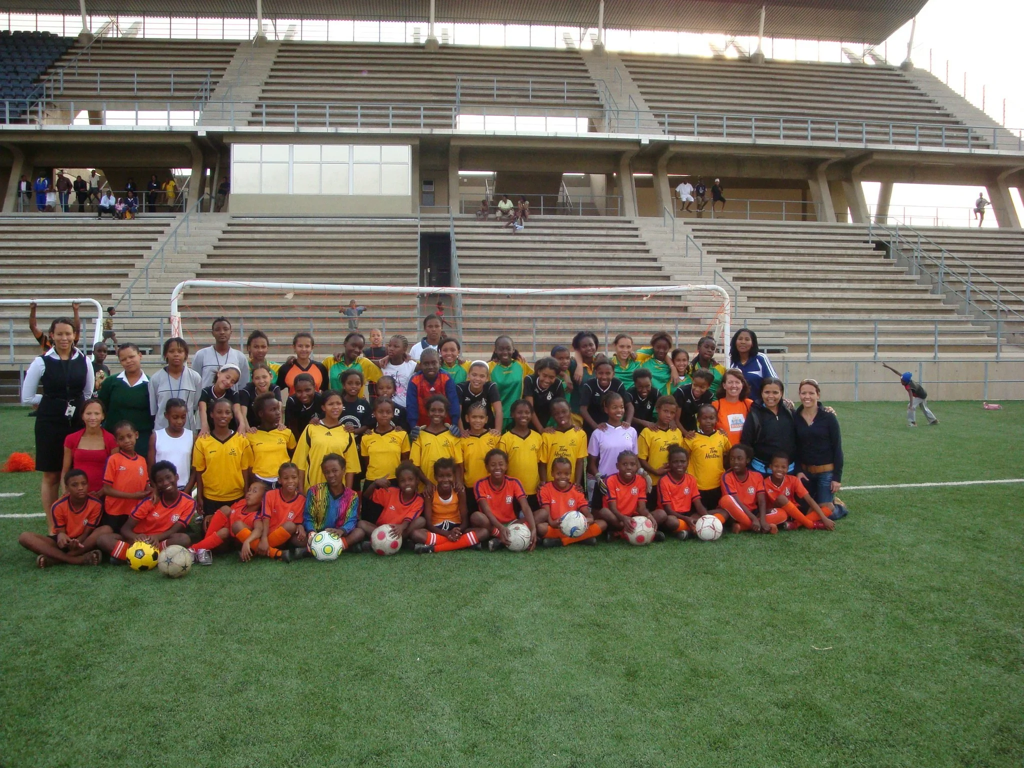 A large group of children and a few adults posing for a photo on a soccer field in front of stadium bleachers. The children are wearing colorful jerseys and are seated or standing with soccer balls in front of them.