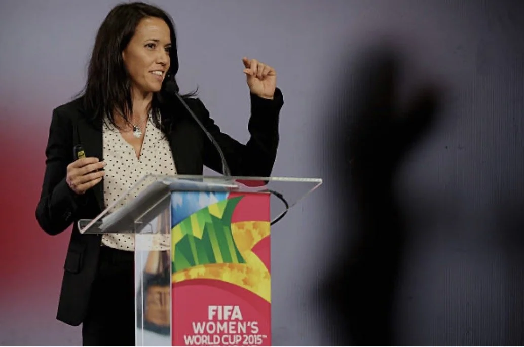 A woman speaking at a podium with a FIFA Women's World Cup 2015 banner, gesturing with her right hand, in front of a gray and black background.