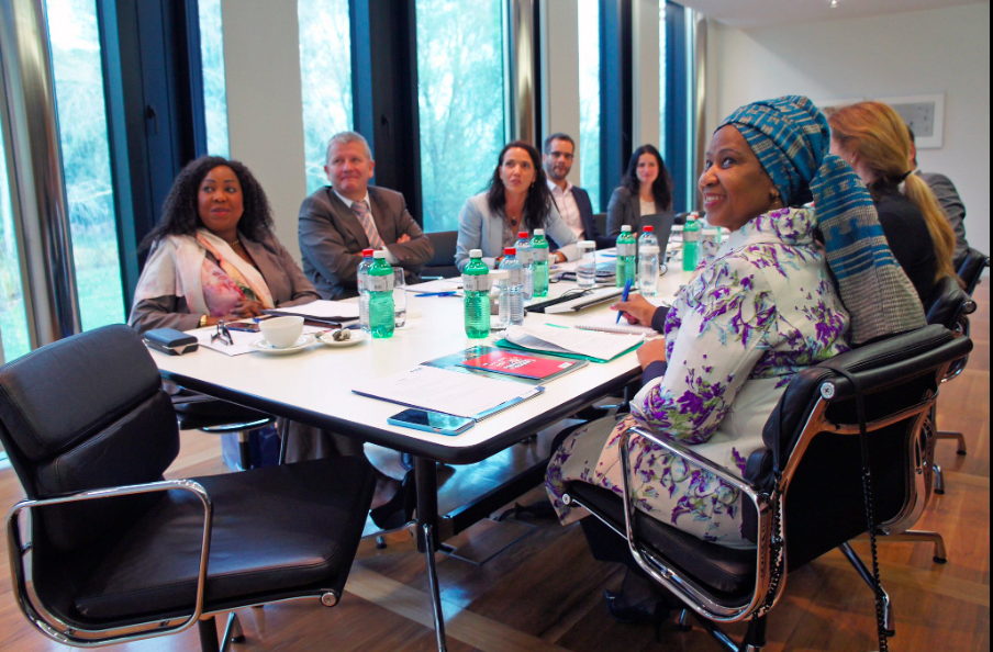 A diverse group of professionals in a business meeting, sitting around a conference table with reports, notebooks, water bottles, and coffee cups, in a well-lit modern office.