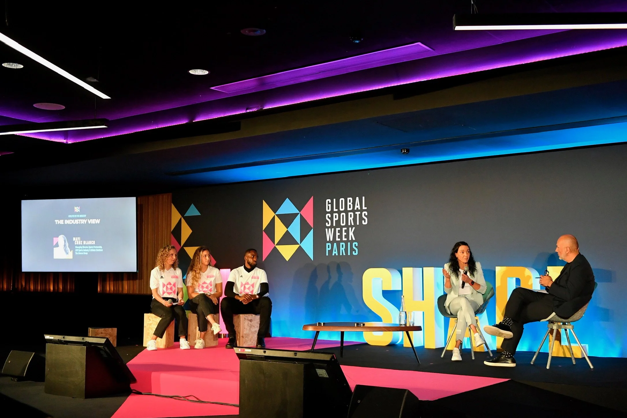 A woman in a light gray suit and white sneakers standing in front of a step-and-repeat banner with logos for Adidas, French Sport, and Global Sports Week Paris.