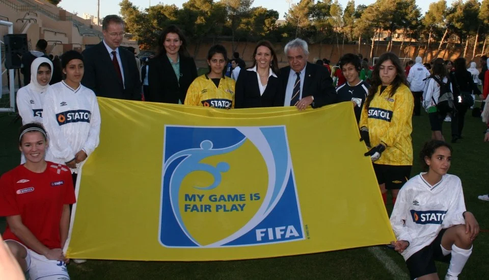 Group of young female soccer players and adults holding a yellow banner with the FIFA logo and the text "My game is fair play," on a soccer field during daylight, with other people in the background.