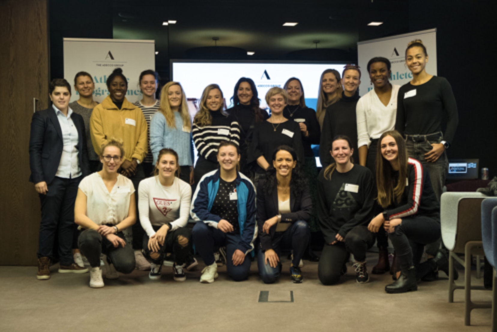 A diverse group of women posing together indoors at a professional event or conference. Some are standing, others kneeling in front. They are smiling and dressed in casual and business casual attire, with a presentation screen and banners in the background.
