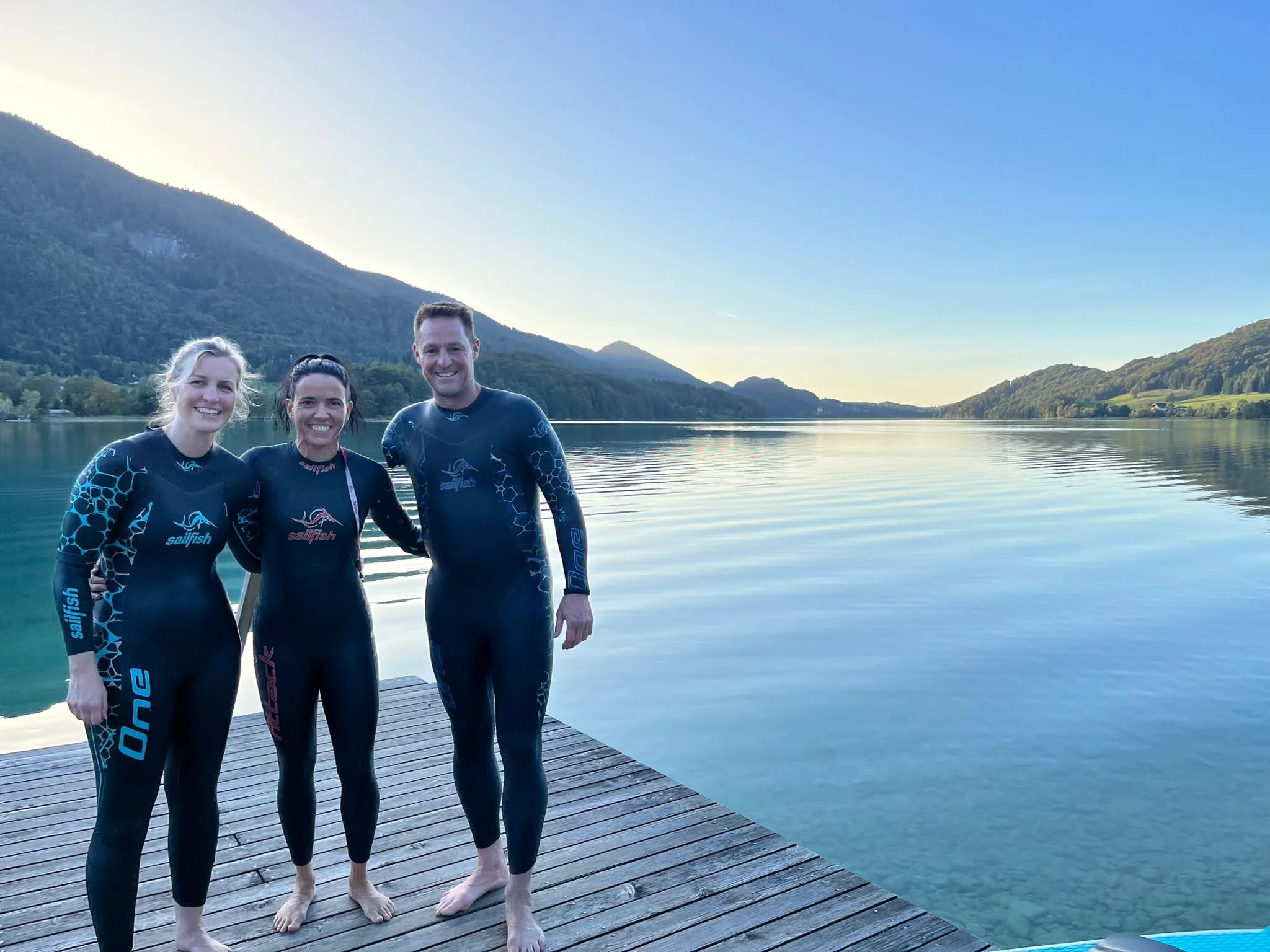 Three people in wetsuits standing on a wooden dock by a lake with mountains in the background.