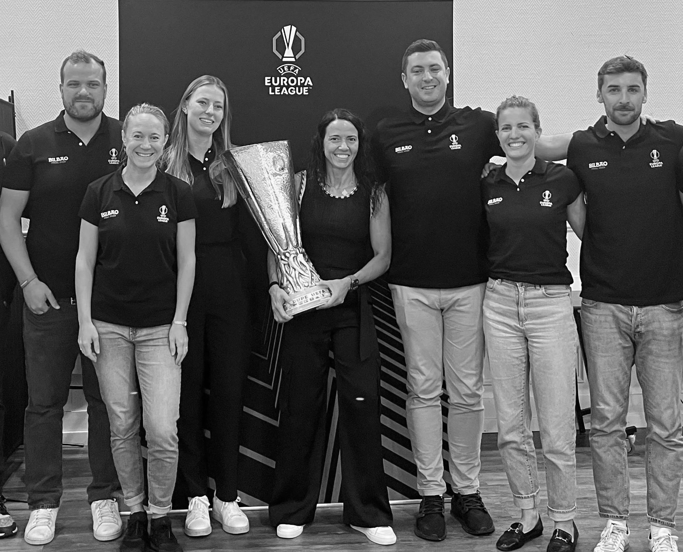 A group of seven people standing together, smiling, with a UEFA Europa League trophy in the center, in front of a backdrop with the UEFA Europa League logo.