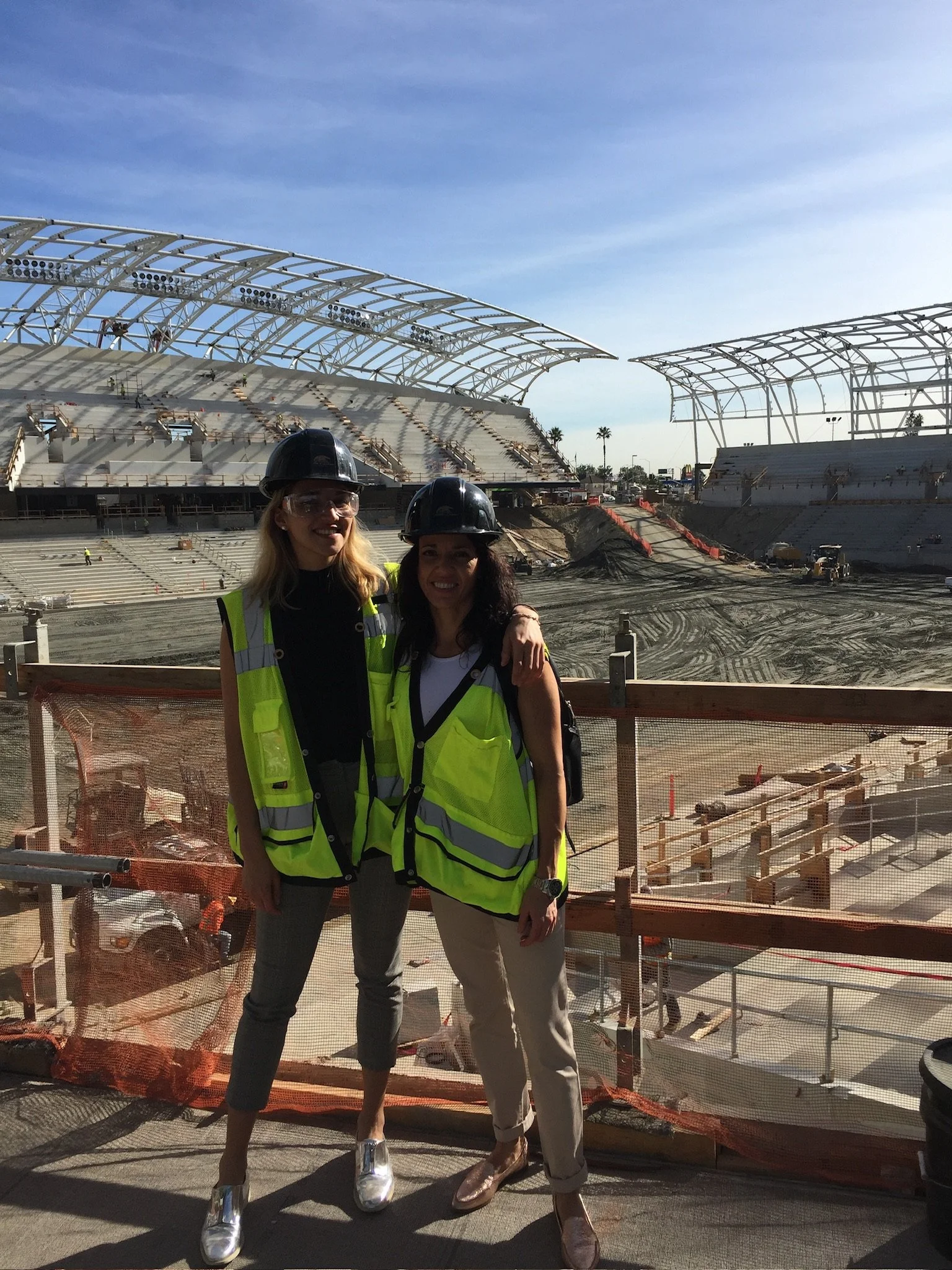 Two women wearing yellow safety vests and construction helmets standing in front of a large stadium under construction, with part of the structure visible behind them.
