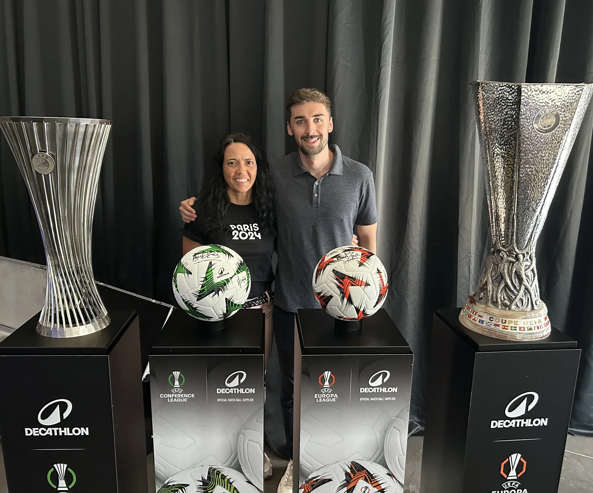 Two people standing behind display cabinets with UEFA and Decathlon branding, holding signed UEFA soccer balls, with UEFA trophies in the background.