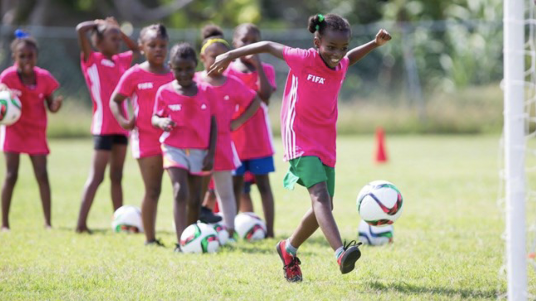 Young girl kicking a soccer ball during practice, with several other girls waiting in line behind her on a grassy field.