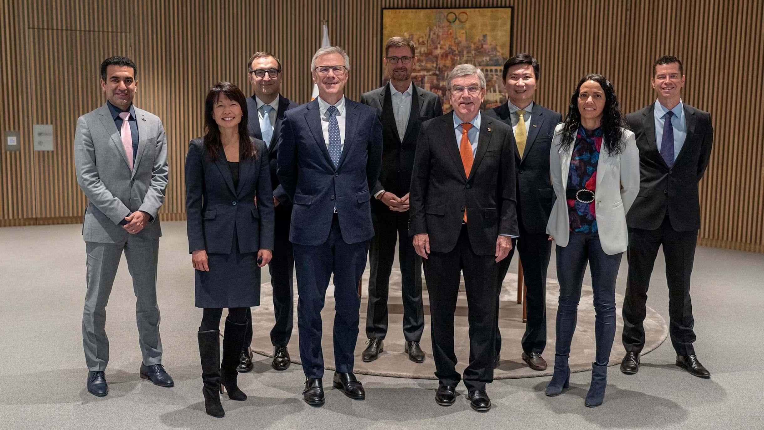 A group of ten professionally dressed individuals standing together indoors with wooden panel walls and a framed painting in the background.