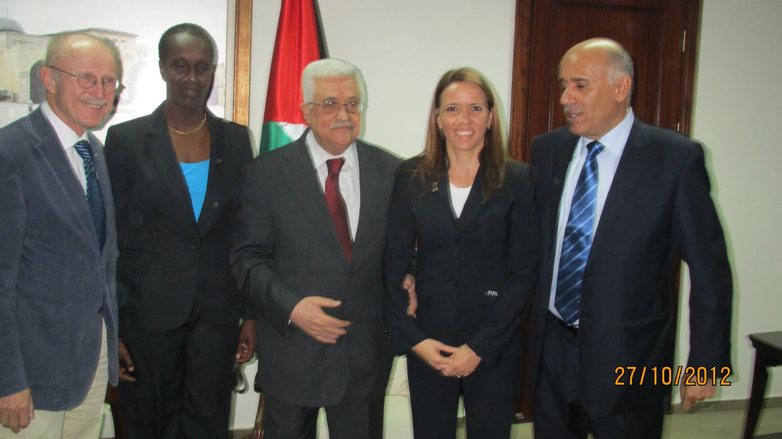 Group of five individuals standing together indoors with a flag behind them, dressed in formal business attire, smiling and posing for the photo.