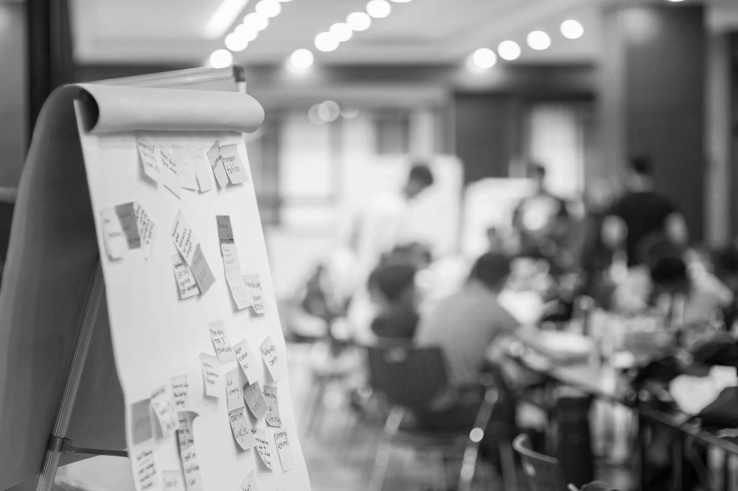 Black and white photo of a flip chart with sticky notes attached to it. The notes contain handwriting and appear to be part of a brainstorming or planning session. In the background, blurred figures are seated around a table in what looks like a conference room.