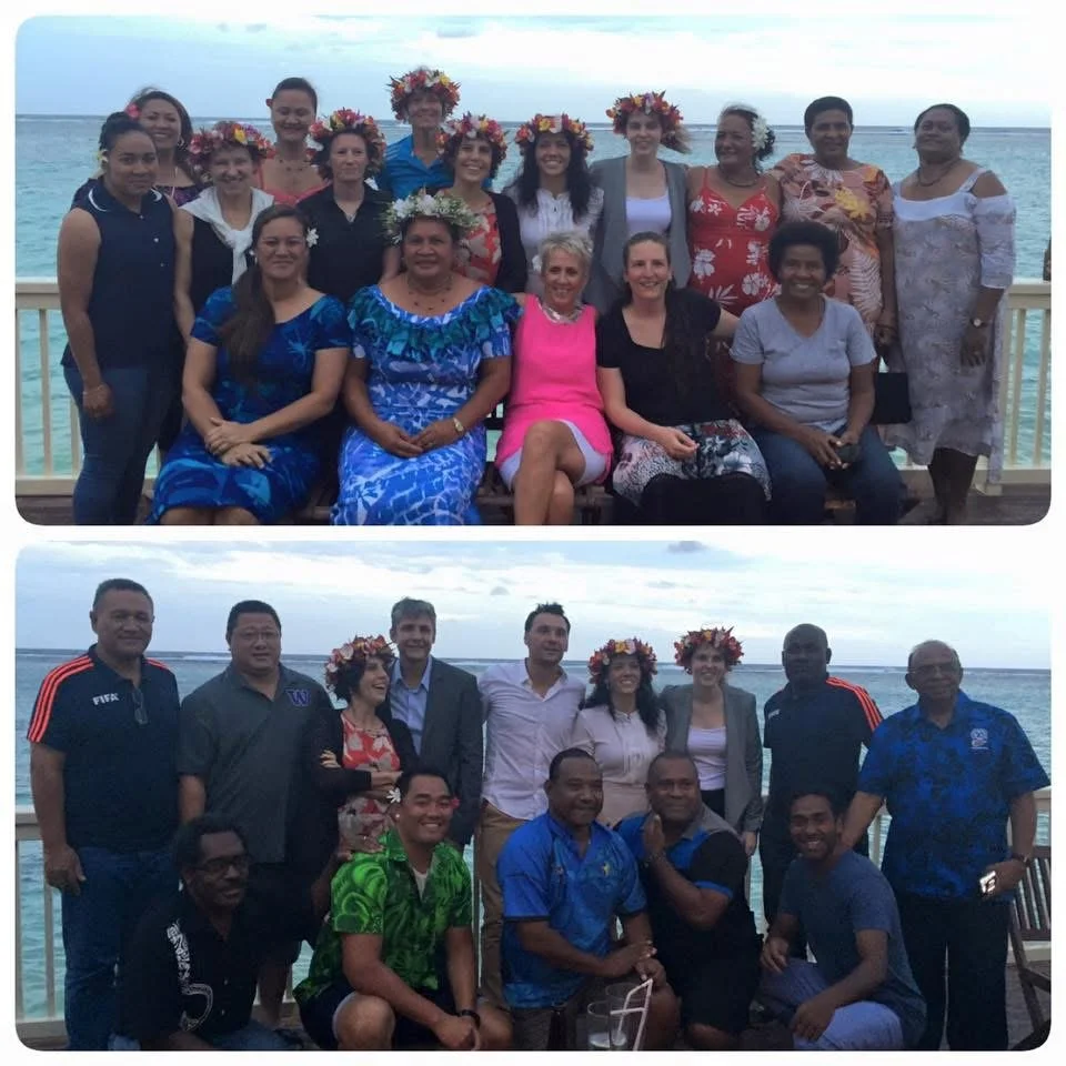 Group of people at a beachside gathering, some wearing floral wreaths, smiling and posing for a photo.