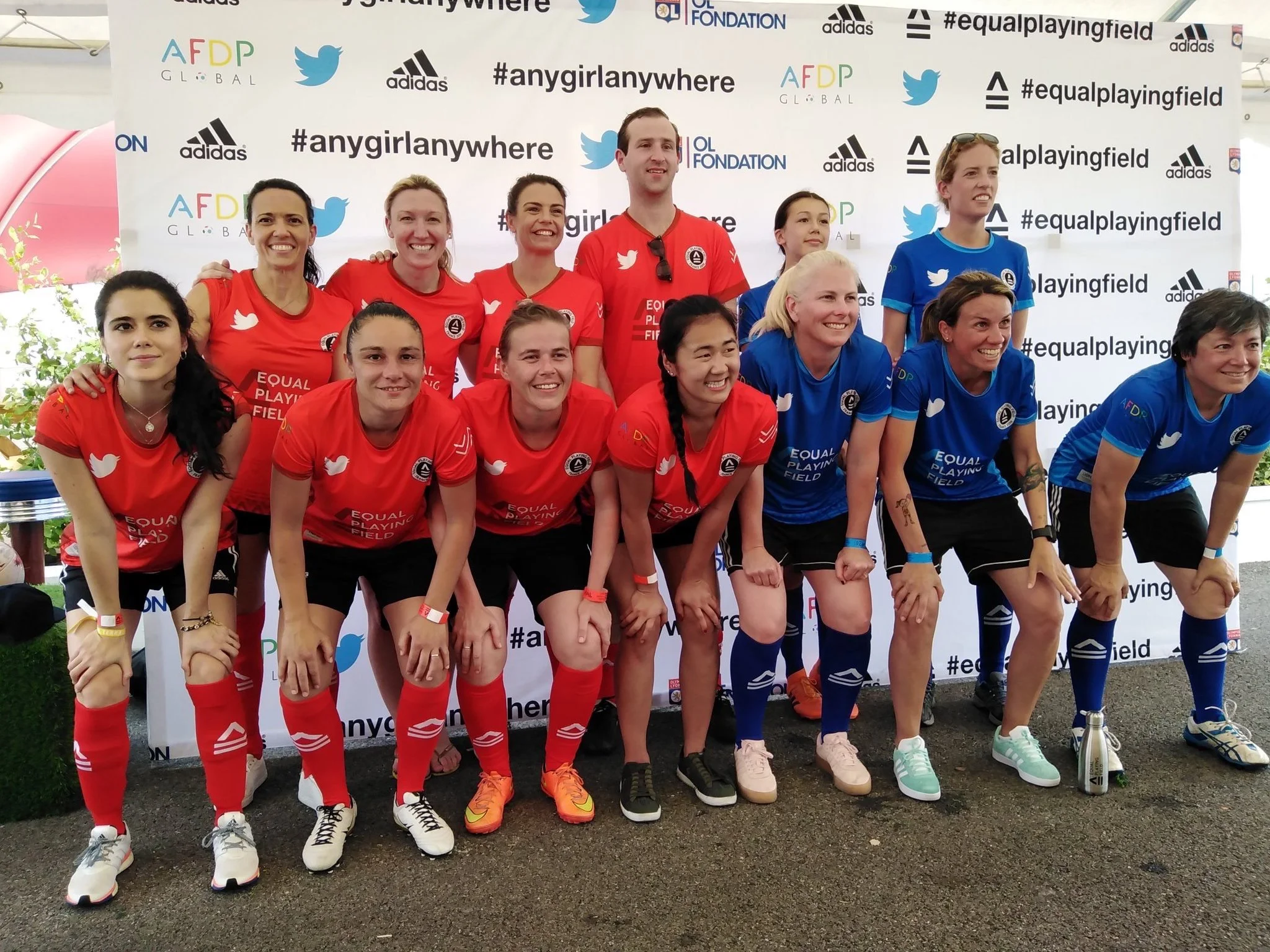A group of women and one man, dressed in red and blue sports uniforms, posing together in front of a branded backdrop at an outdoor event. The women are smiling and some are kneeling in the front row, while others are standing behind them.