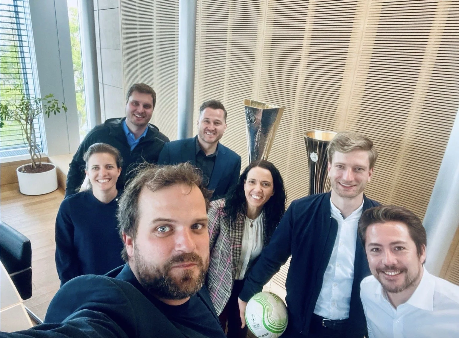 A group of eight people, five men and three women, taking a selfie in a modern office or conference room with wooden wall panels, large windows, potted plant, and trophies in the background. Some individuals are holding a volleyball.