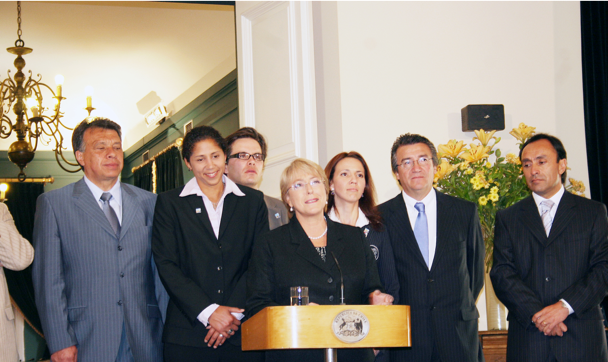 Group of six people standing behind a podium, with a woman in the center speaking, indoors with chandelier and floral decor.