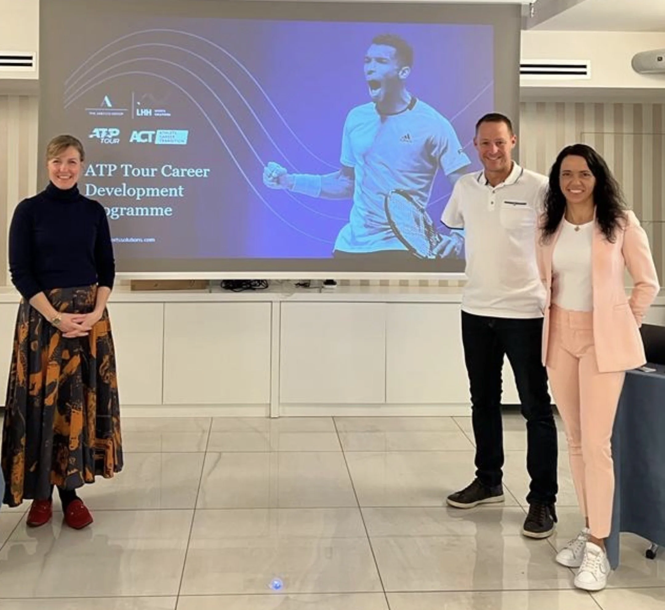 Four people standing in front of a large projection screen displaying logos and text related to an ATP tennis career development program. The five individuals are smiling, with one woman on the left wearing a black turtleneck and patterned skirt, a man second from the right in a white shirt, and a woman on the far right in a pink suit. The person on the projection screen is a male tennis player wearing a light blue Adidas shirt, celebrating with his fist clenched.