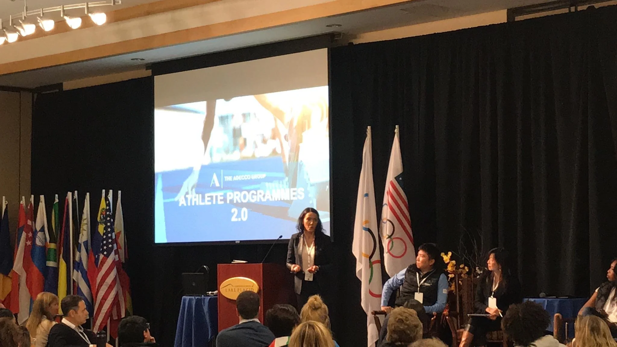 A conference room with a large screen displaying a presentation titled 'Athlete Programs 2.0' and the logo of The Adecco Group. Several flags from different countries are on display, and people are seated in the audience with some individuals standing or on stage.