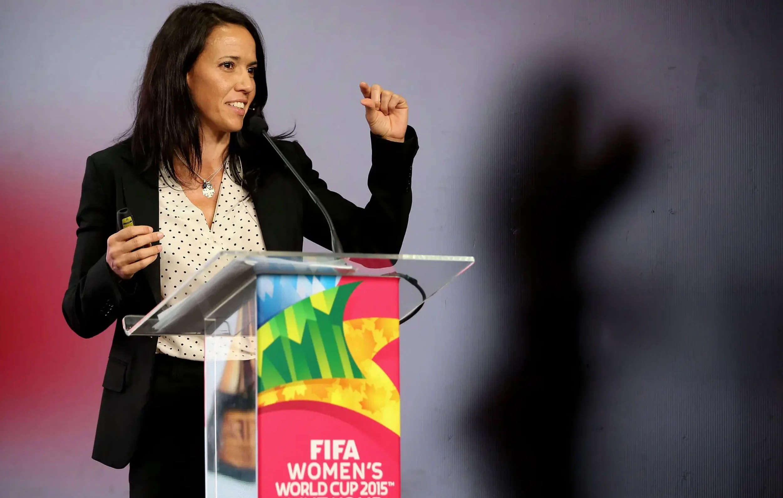 Mayi Cruz Blanco, wearing a black blazer and a white blouse with black polka dots, is standing at a podium with a FIFA Women's World Cup 2015 banner. She is speaking into a microphone and gesturing with her right hand.