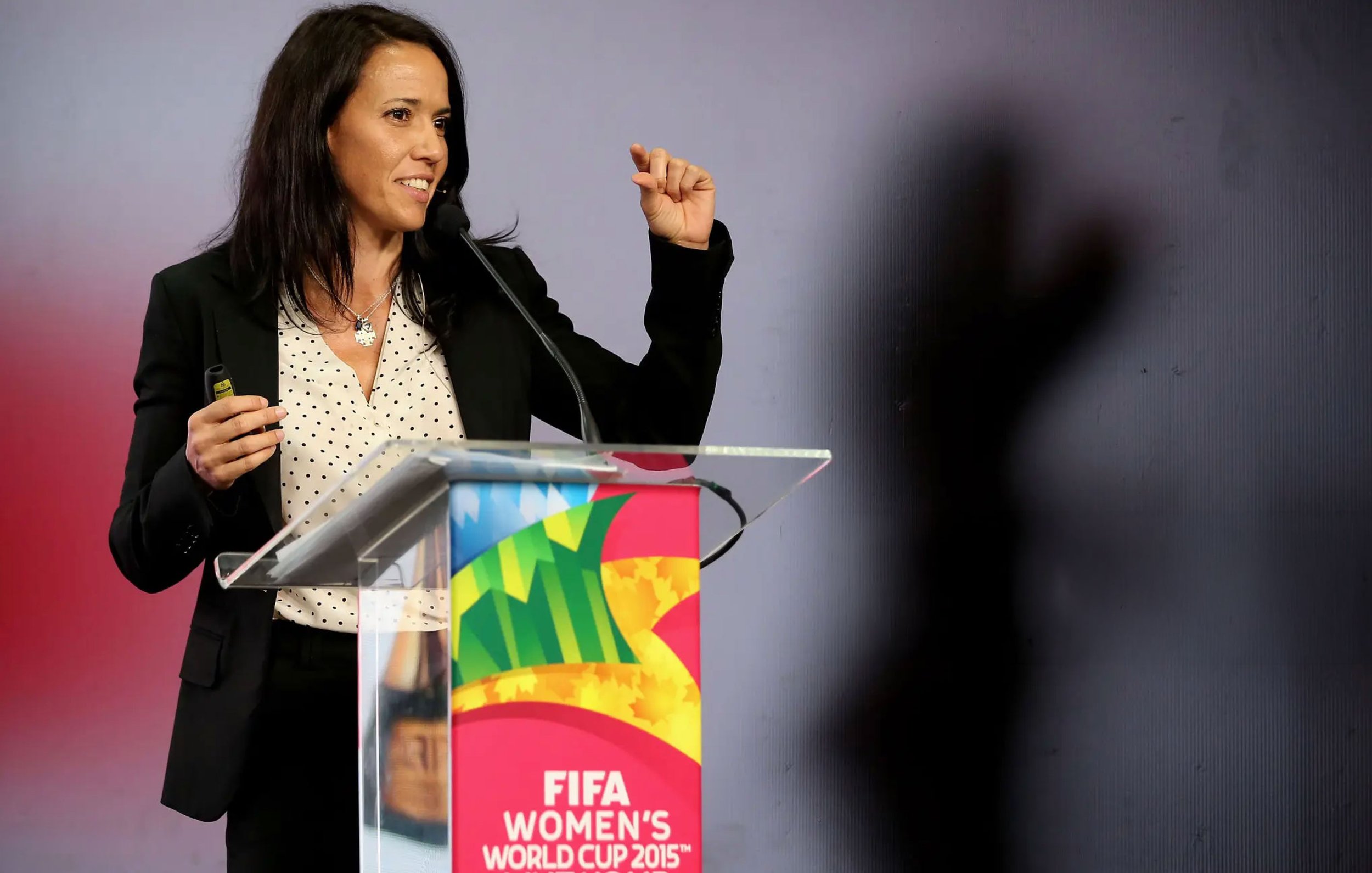 Mayi Cruz Blanco, wearing a black blazer and a white blouse with black polka dots, is standing at a podium with a FIFA Women's World Cup 2015 banner. She is speaking into a microphone and gesturing with her right hand.