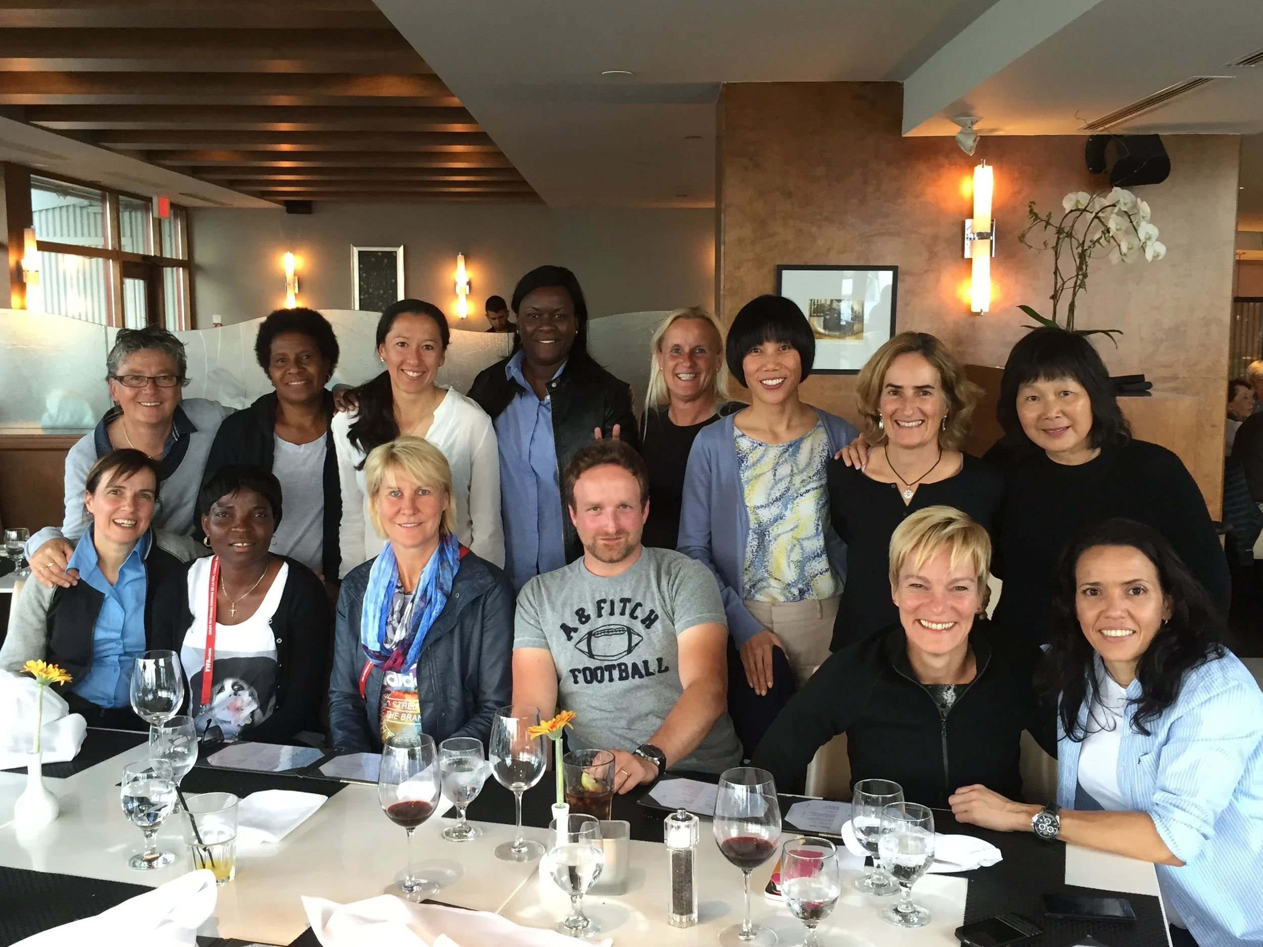Group of smiling people gathered at a restaurant table with wine glasses, plates, and flowers.