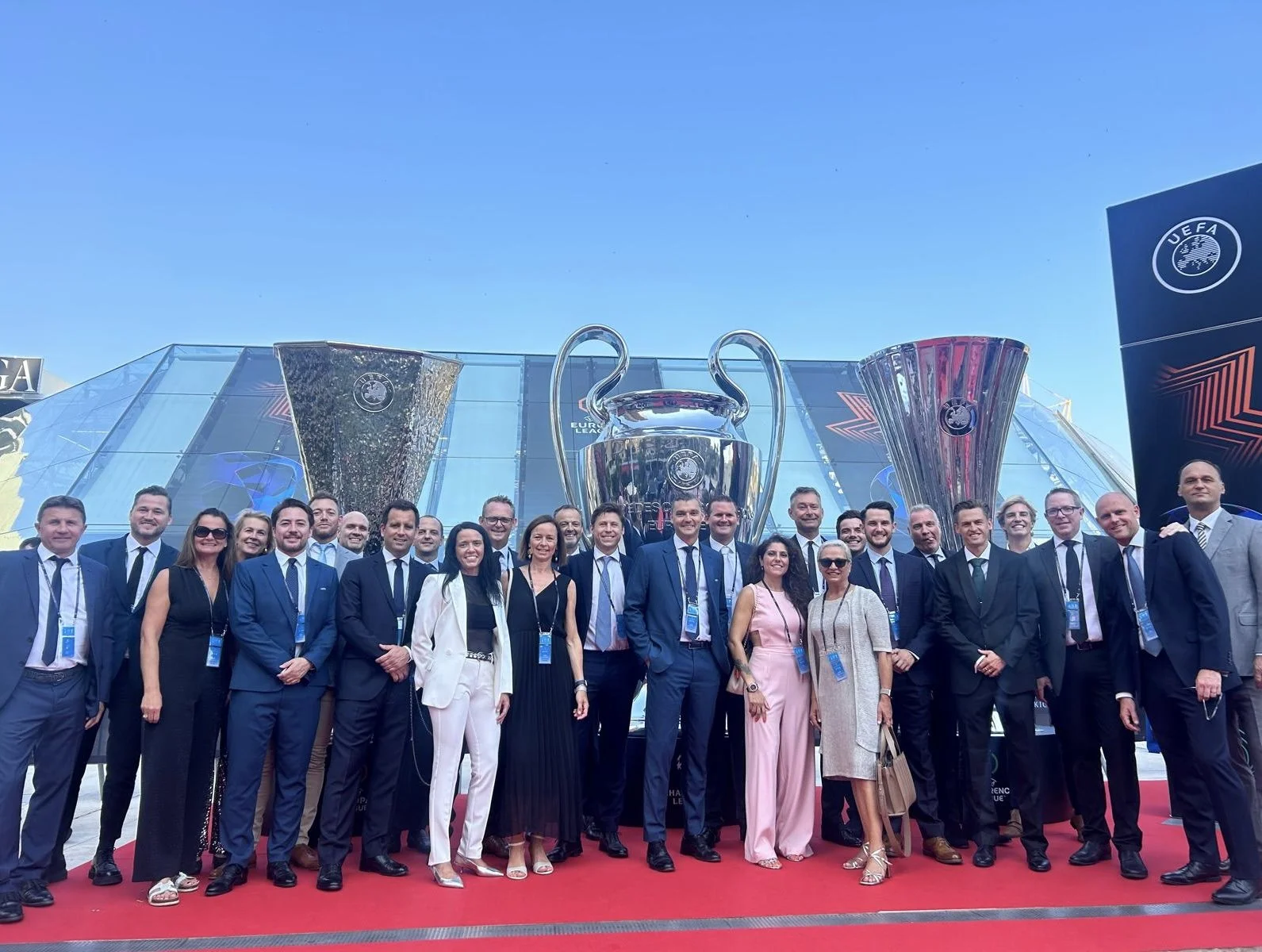 A large group of professionally dressed people standing on a red carpet in front of large UEFA trophies, including the Champions League trophy, with a modern glass building and blue sky in the background.