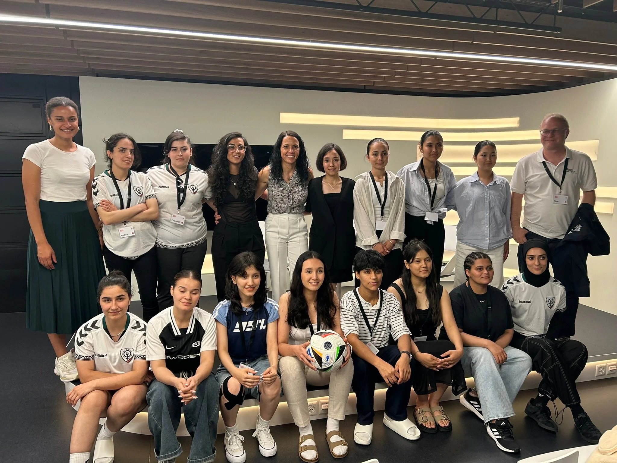 Group of women and a man posing for a photo in what appears to be a conference or event room, some women wearing sports jerseys, one woman holding a soccer ball.
