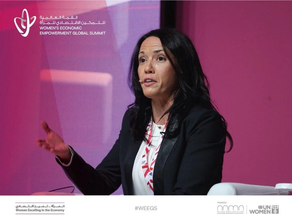 A woman speaking at the Women's Economic Empowerment Global Summit with a pink backdrop and summit branding in Arabic and English.