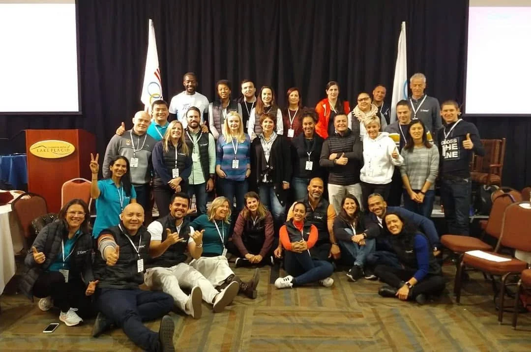 Group photo of around 25 diverse people at a conference or event, some making peace signs or thumbs up, standing and sitting in front of a stage with black curtains and two screens, at a venue called Lake Placid.