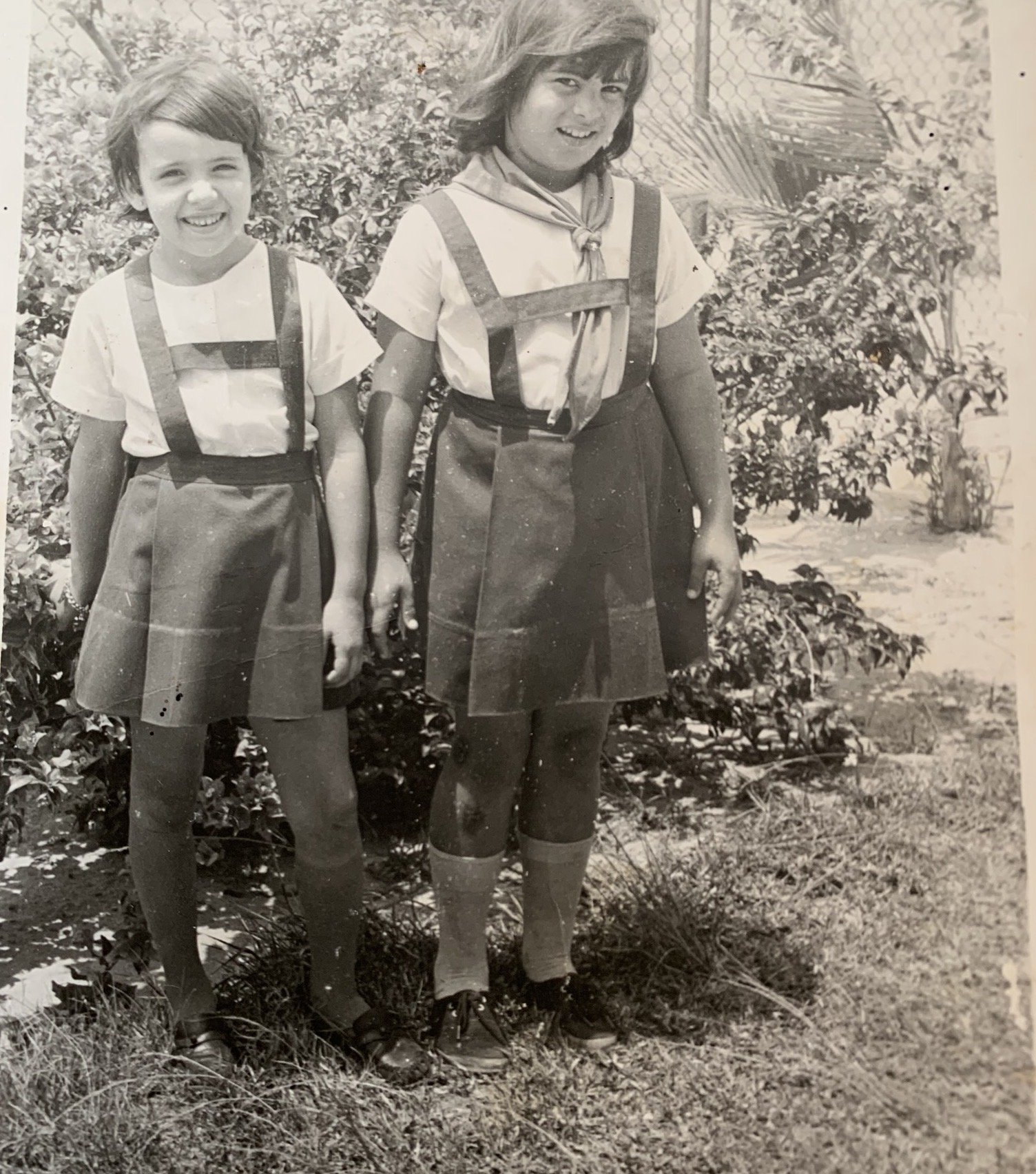 Two girls wearing scout uniforms, including neckerchiefs and shorts, standing outdoors near bushes and plants, smiling at the camera in this black-and-white photograph.