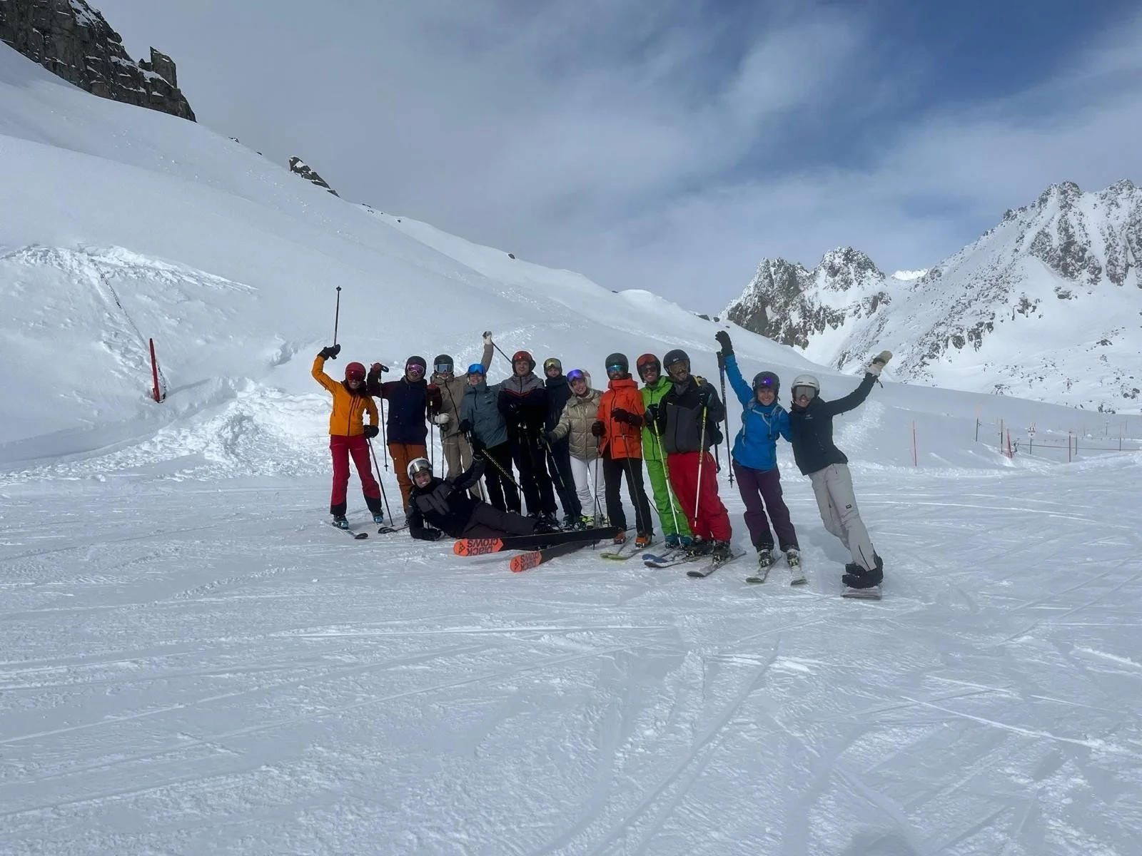 Group of people in winter skiing gear celebrating on snowy mountain slope with ski equipment, mountains, and cloudy sky in the background.