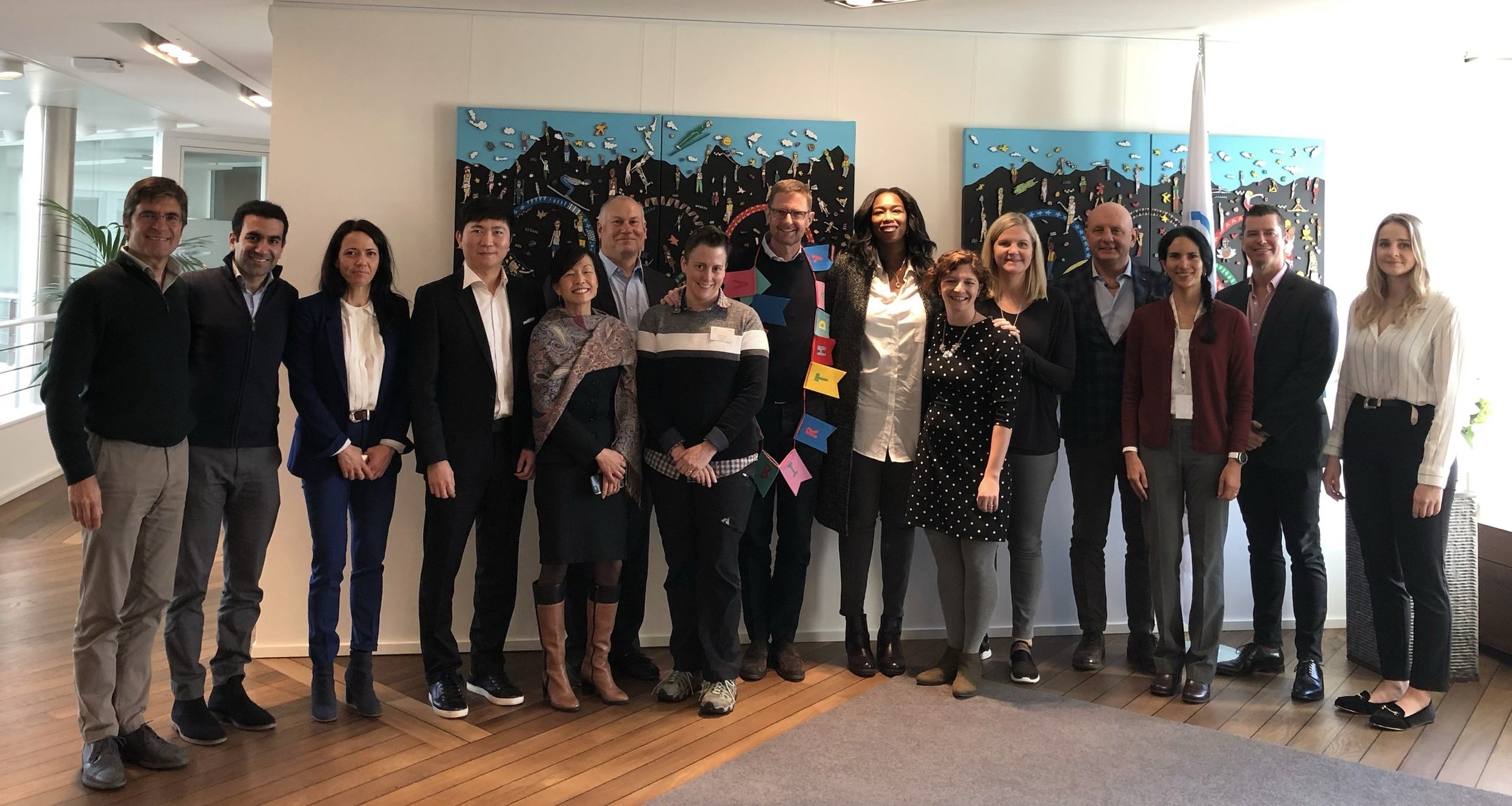 A diverse group of sixteen people standing together indoors, smiling for a photo with a colorful art piece in the background.