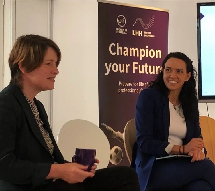 Two women in business attire sitting on a stage during a panel discussion, with a banner behind them that reads "Champion your Future" and features logos for Women in Football and Sports Solutions.