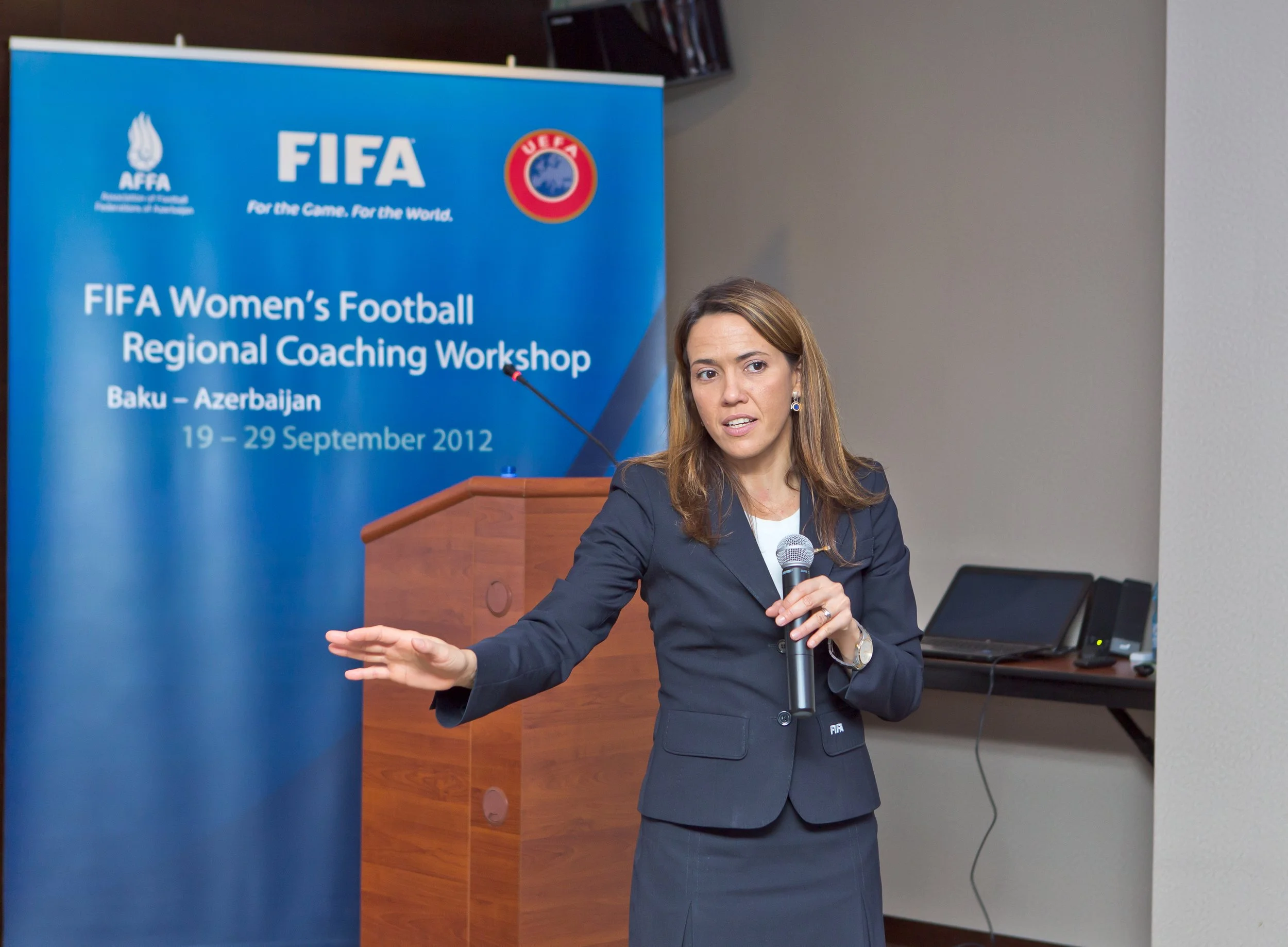 A woman with brown hair, wearing a dark blue suit, speaking into a microphone at a conference. Behind her, there is a blue banner with logos and text about a FIFA Women's Football Regional Coaching Workshop held in Baku, Azerbaijan, from September 19 to 29, 2012.