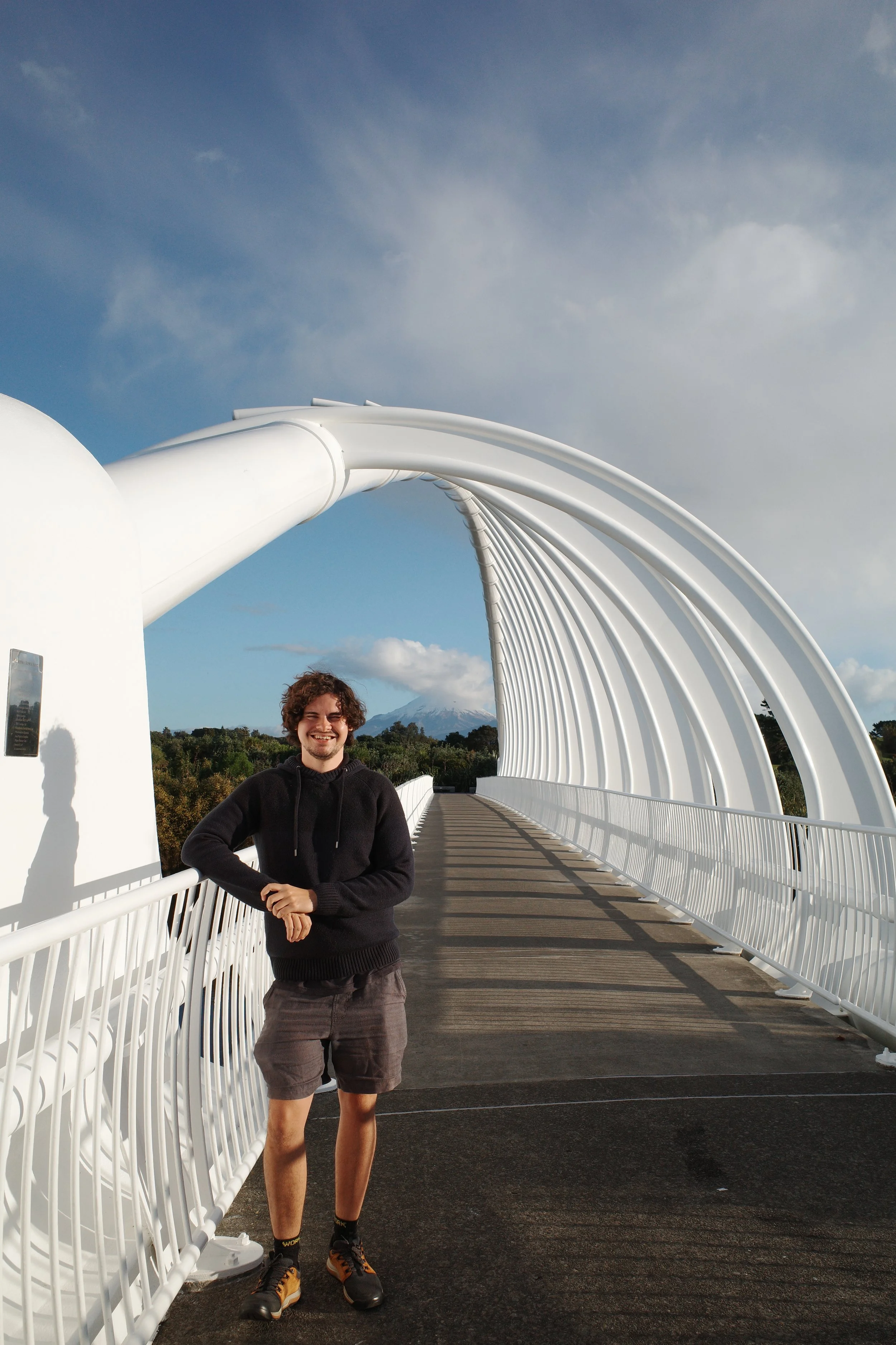 A young man with curly hair smiling and wearing a black hoodie, shorts, and hiking shoes, standing on a modern white arched pedestrian bridge with a scenic background including trees, mountains, and a partly cloudy sky.