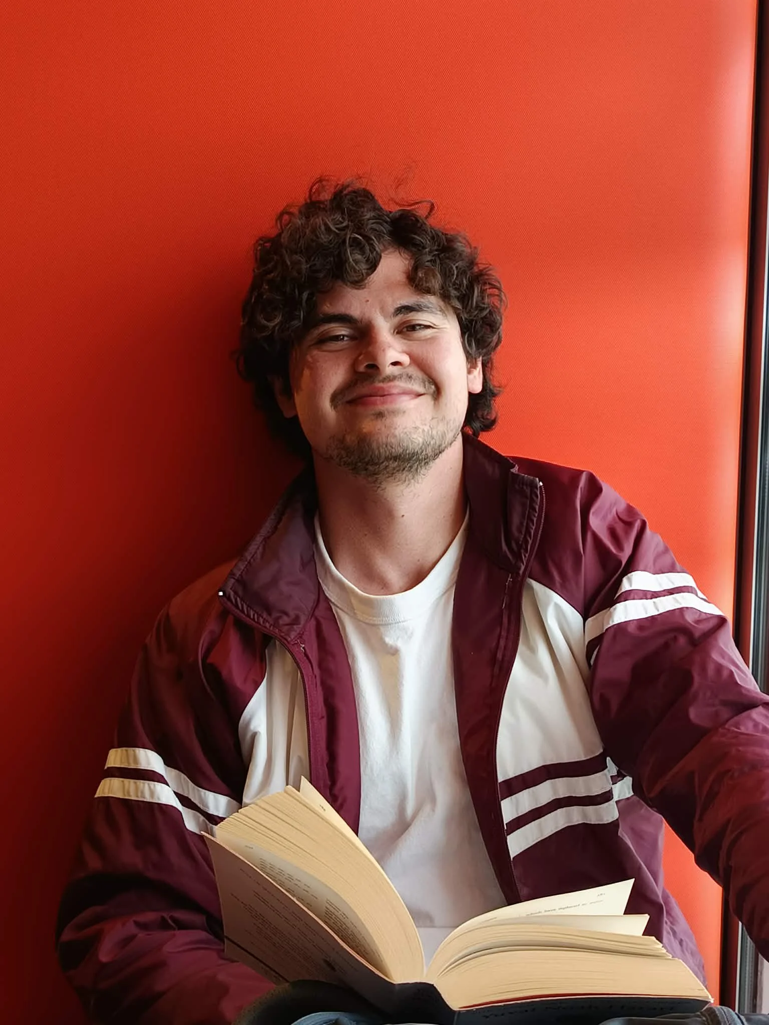 A young man with curly hair, light skin, and a beard, smiling while reading a book, sitting against a vibrant orange wall.