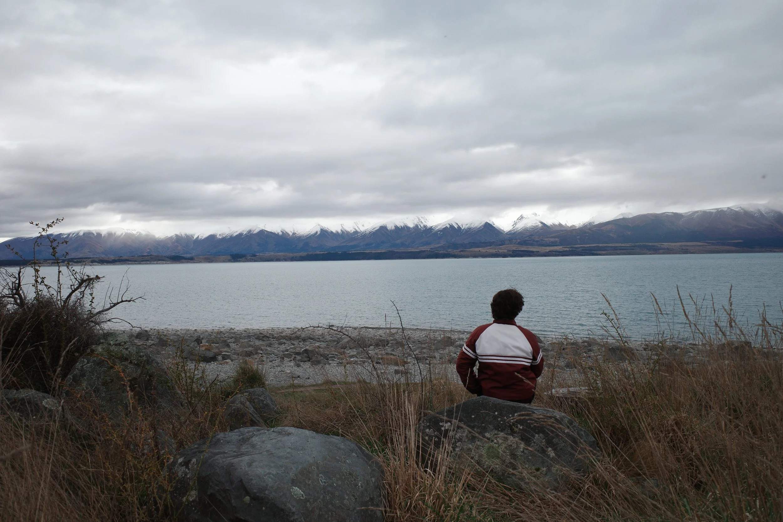 Person sitting on rocky shore facing a lake with snow-capped mountains under cloudy sky.