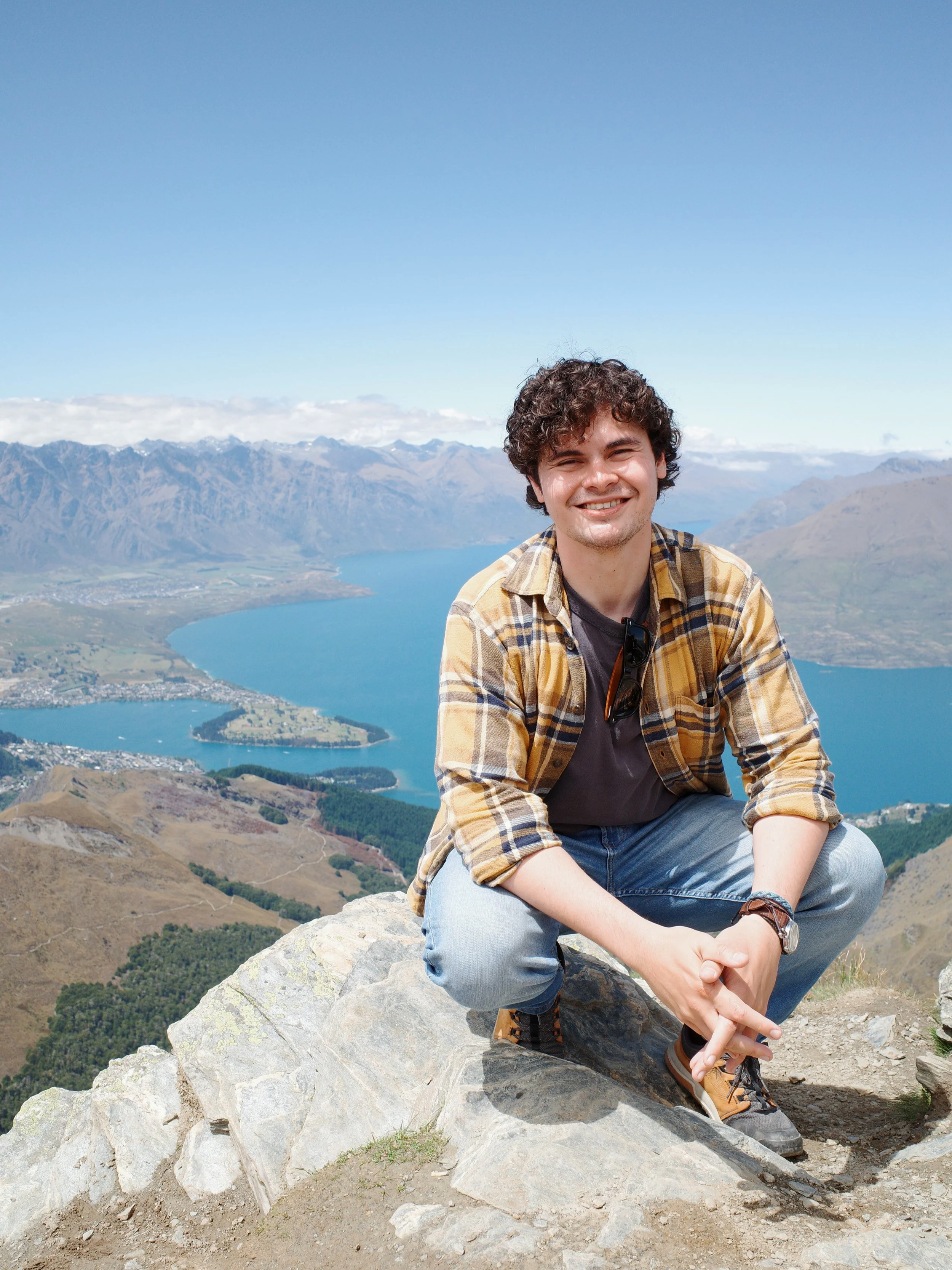 A young man with dark curly hair, wearing a plaid shirt over a gray t-shirt, sitting on a rock on a mountain trail with a scenic view of a lake, mountains, and partly cloudy sky in the background.