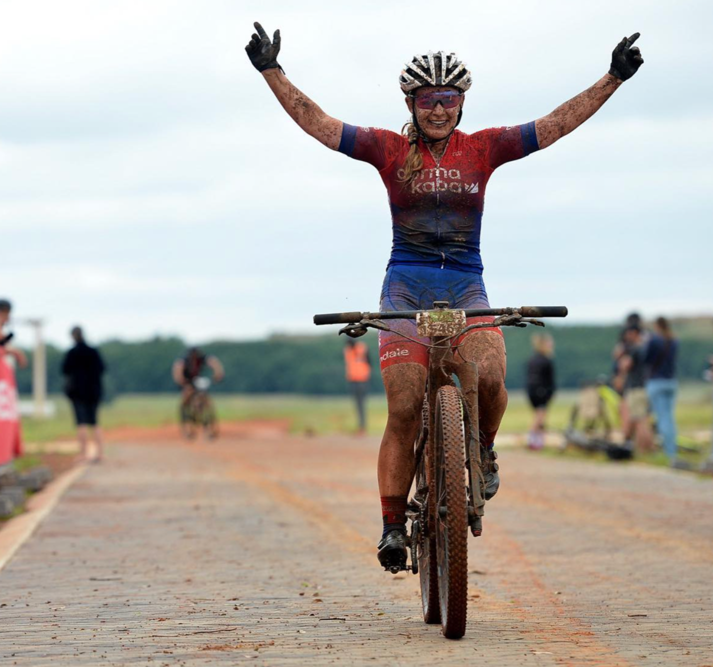 Amy Wakefield rides a muddy mountain bike on a dirt track, raising her arms in celebration.