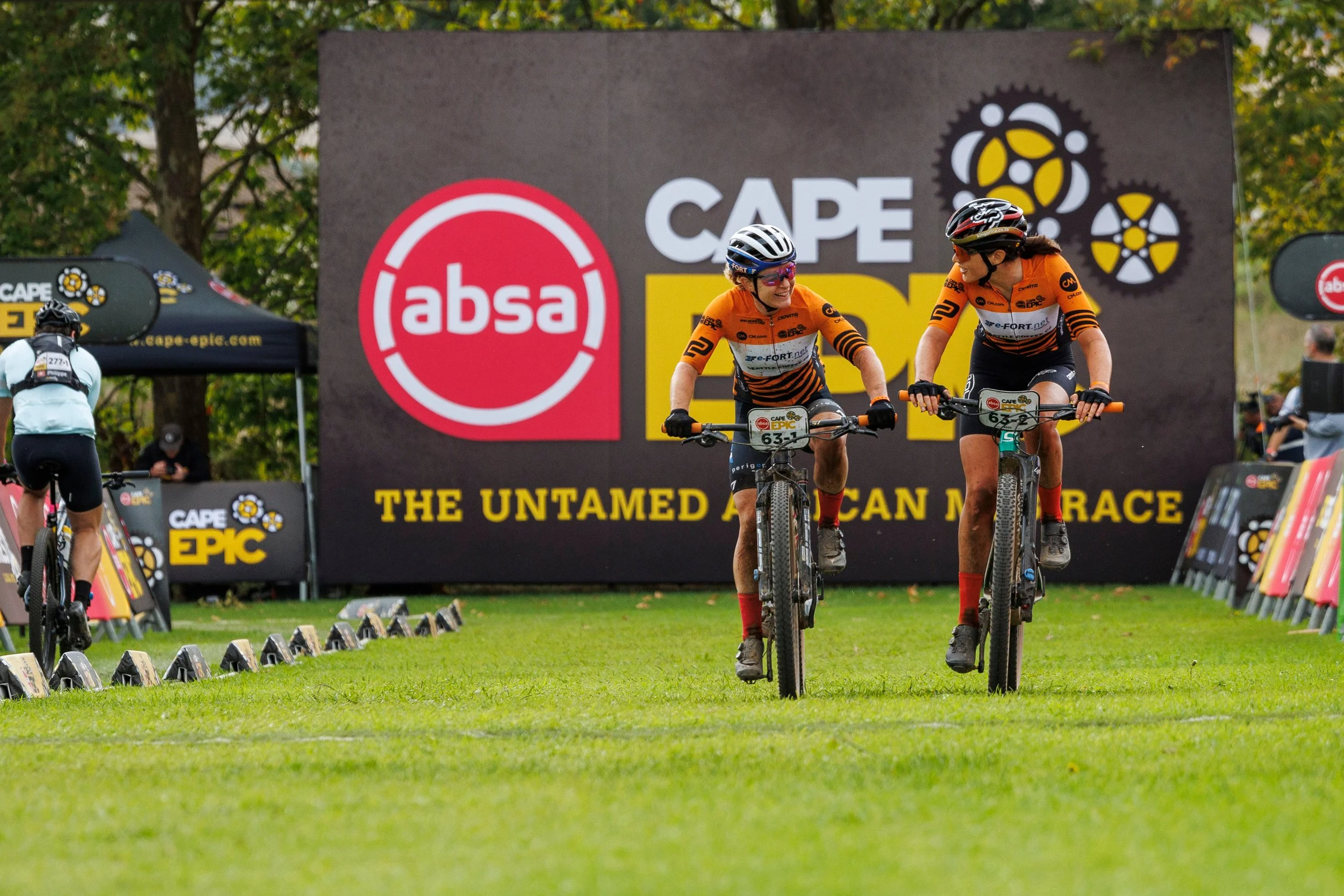 Two female mountain bikers wearing orange and black jerseys and helmets, smiling and talking as they navigate a race course with a large sign that reads "Cape Epic" and bears the logo of ABSA bank. There are other participants and spectators in the background, with trees and race equipment visible.