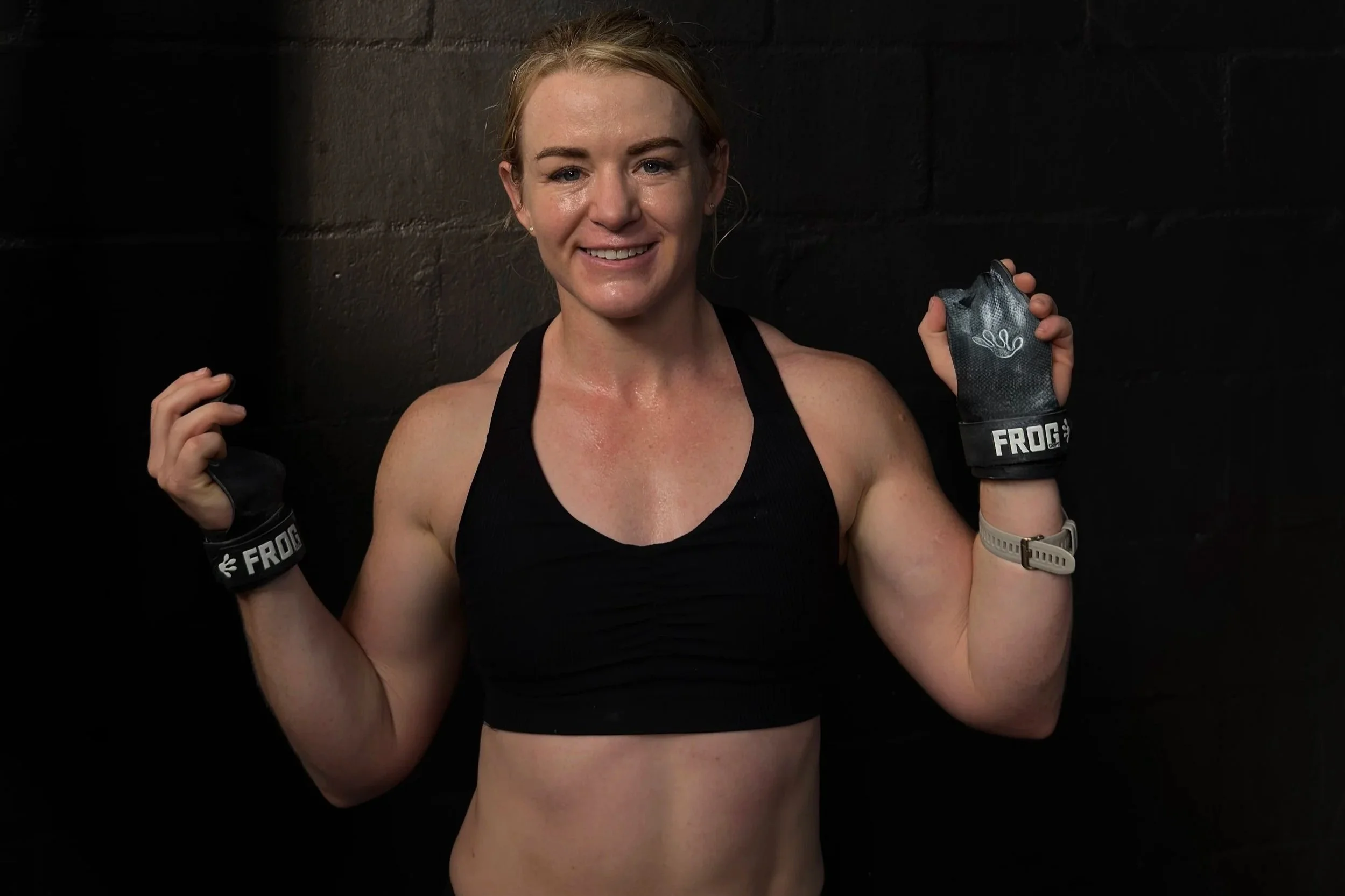 Amy Wakefield, wearing a black sports top, black gloves with 'FROG' written on them, holding her fists raised, standing against a dark wall after a workout.