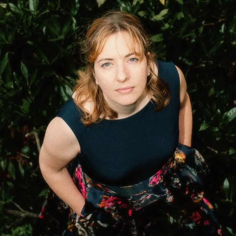 Junge Frau mit lockigem, hellbraunem Haar und blauen Augen, trägt ein dunkelblaues Top und eine bickfarbe Bluse mit Blumen, steht vor dunklen grünen Blättern.