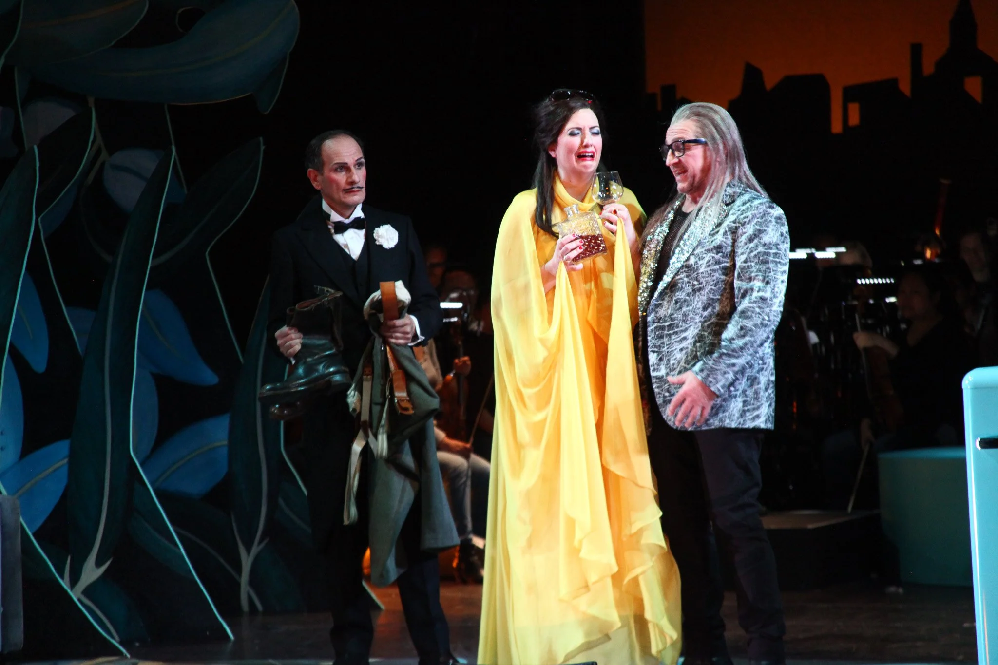 Drei Personen auf einer Bühne, zwei Männer und eine Frau, die in einer Theaterproduktion gekleidet sind. Die Frau trägt ein großes, gelbes Kleid und hält ein Weinglas. Der Mann rechts trägt eine auffällige Jacke, die andere trägt einen schwarzen Anzug. Alle scheinen eine emotionale Szene darzustellen.